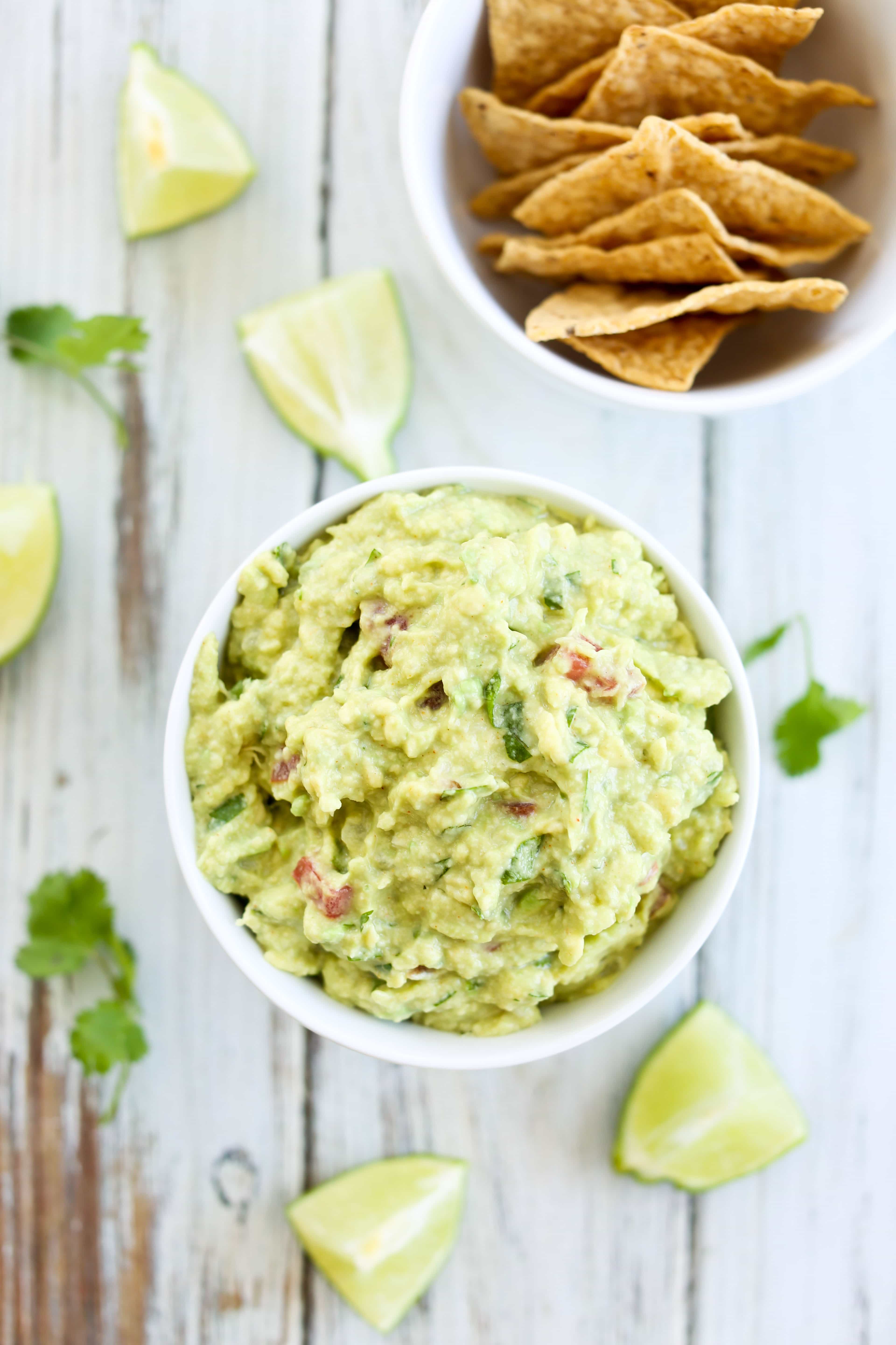 overhead shot of a bowl of the best guacamole with cilantro and lime slices on the table and a bowl of tortilla chips