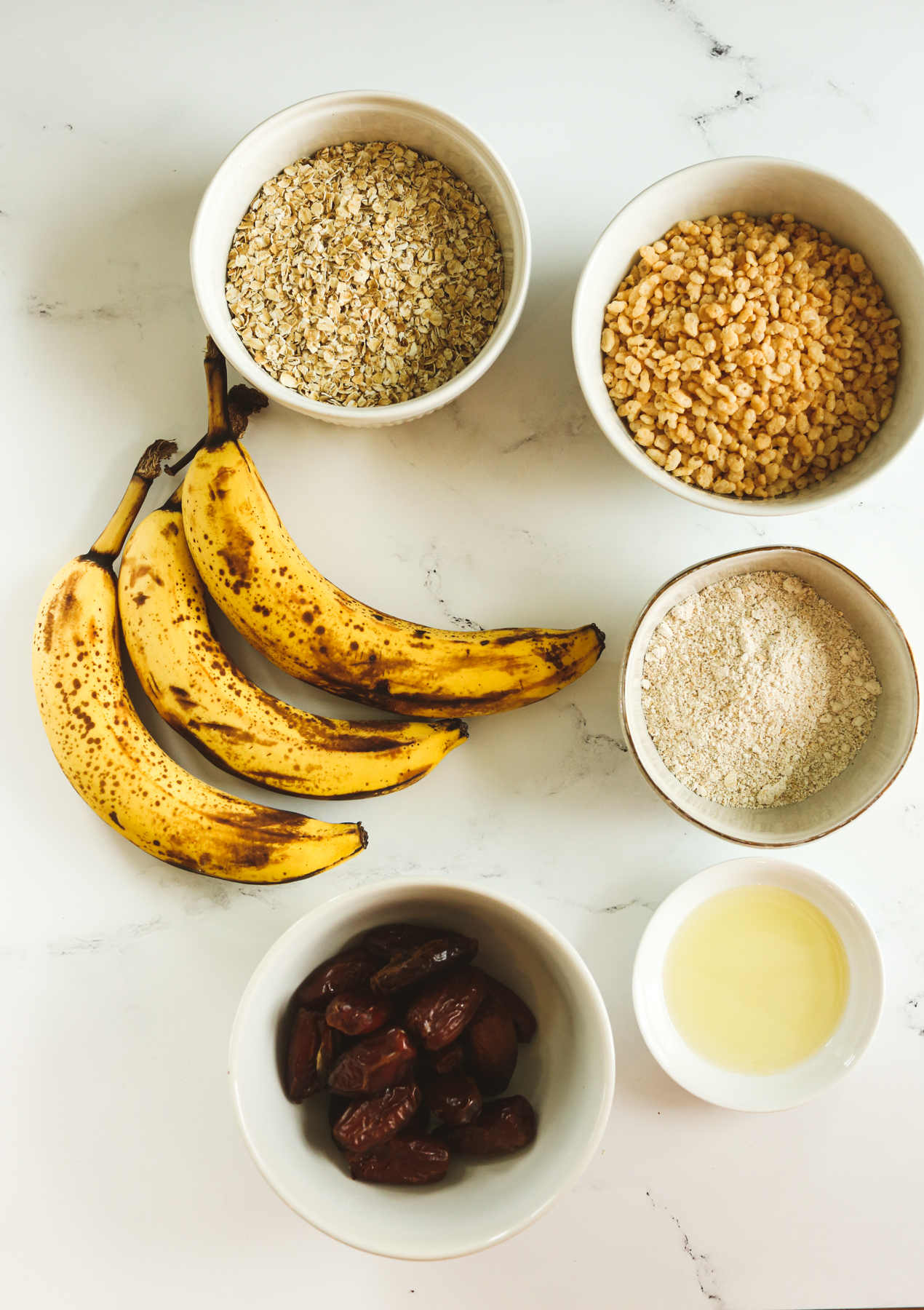 recipe ingredients overhead shot--bananas, bowls with oats, oat flour, dates, brown rice crisp cereal, and oil