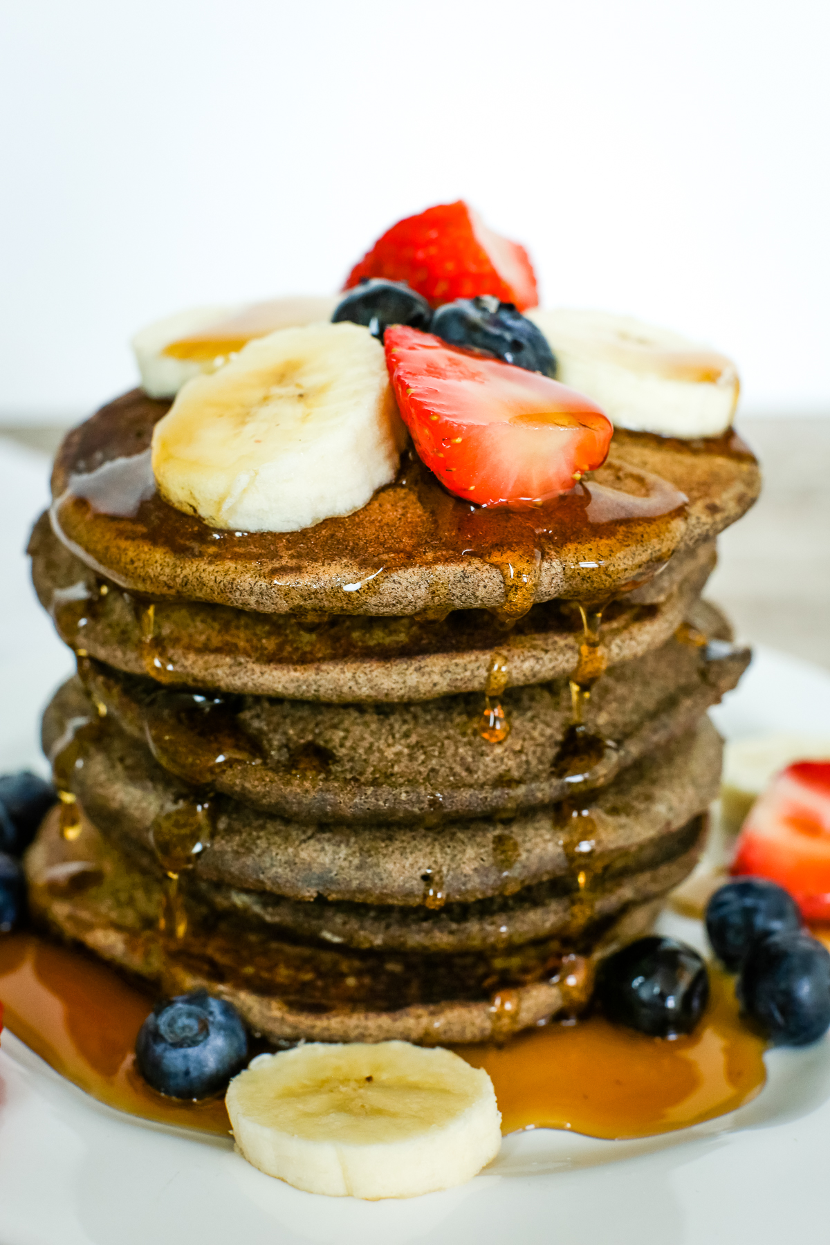 close up view of buckwheat pancakes with strawberries, blueberries, and bananas and syrup dripping down the stack