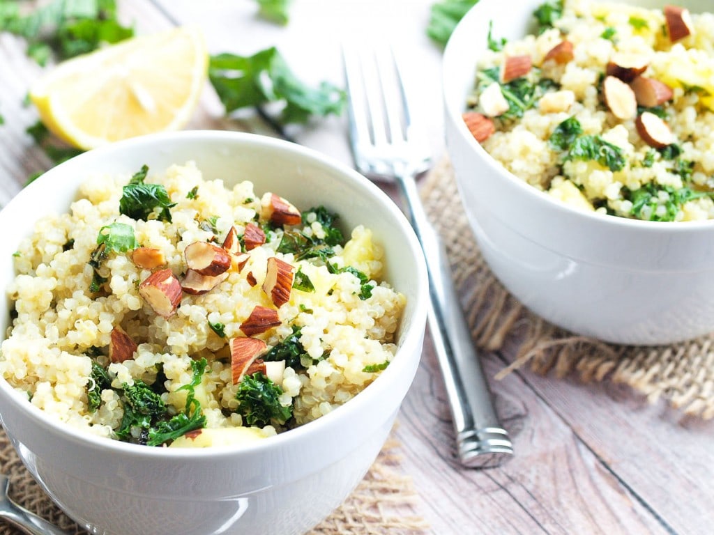 Warm Quinoa Salad with Apples and Kale in two bowls with a lemon on the table and a fork in between the bowls. Almonds are sprinkled on top.