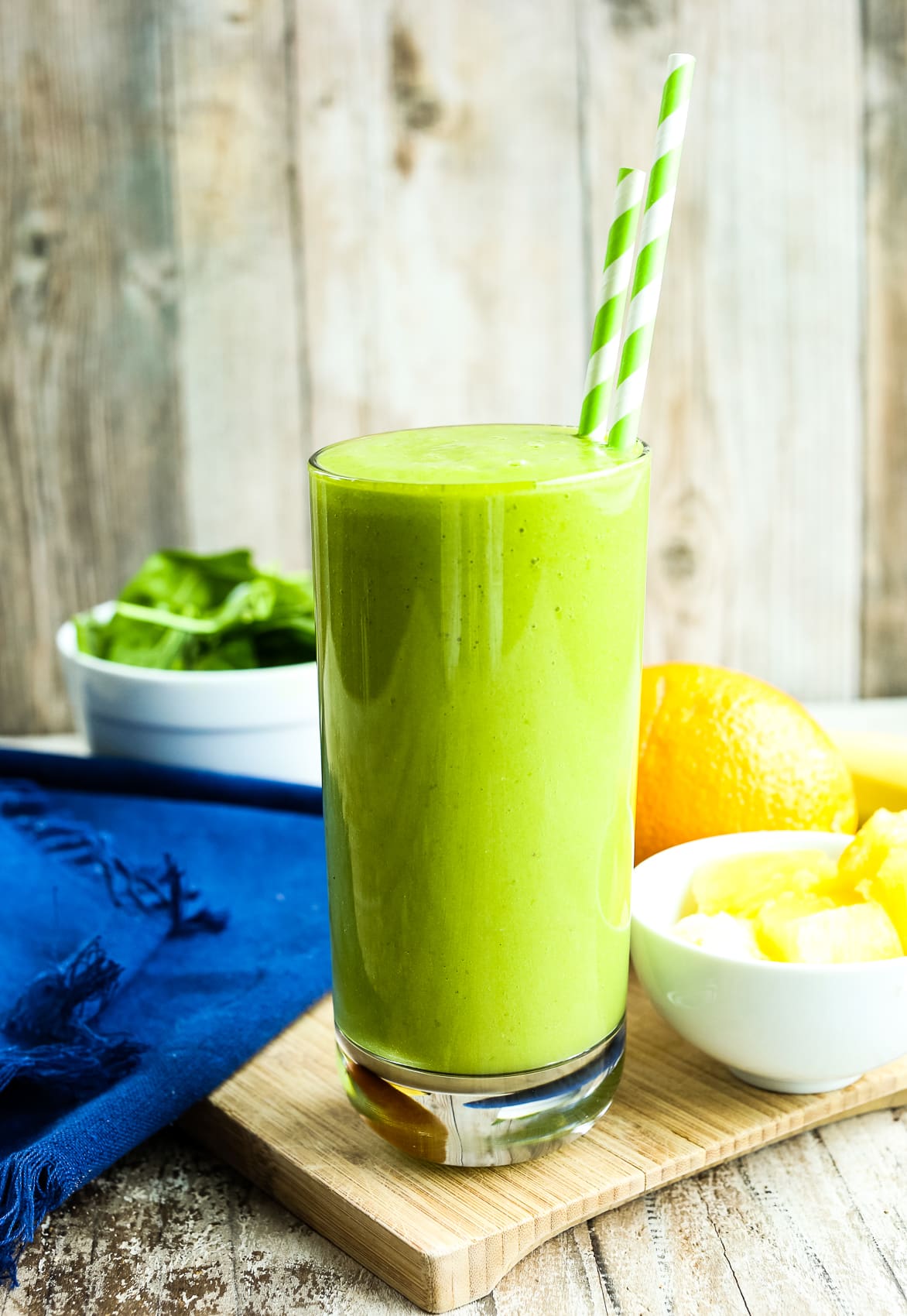 a pineapple spinach smoothie with two green straws in a tall glass with a navy napkin and recipe ingredients in the background