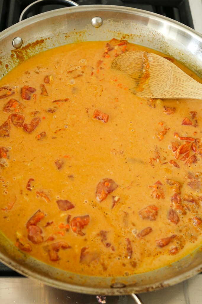 A large skillet with coconut milk, diced tomatoes, minced ginger, and a wooden spoon stirring.