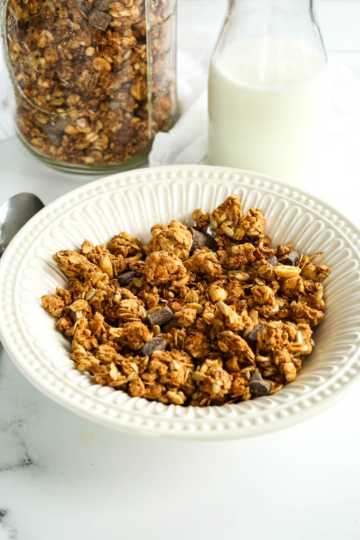 A bowl of cereal with a glass jug of milk in the background and a mason jar filled with the same cereal
