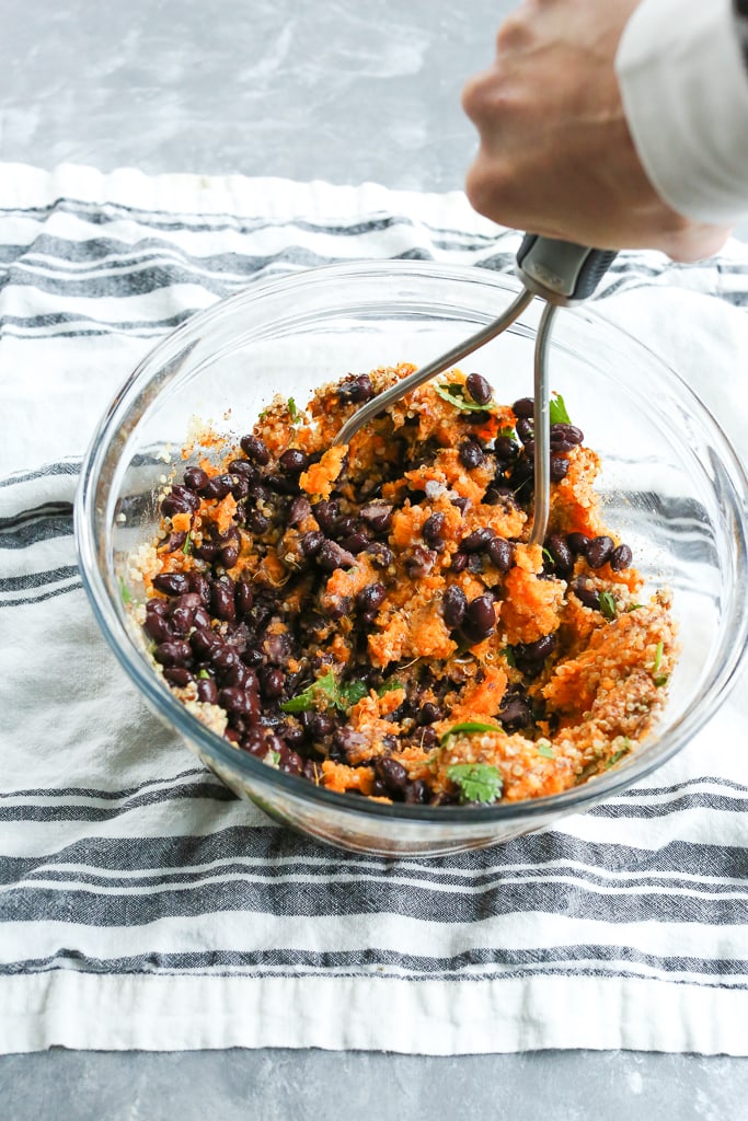 cooked sweet potato, black beans, quinoa, cilantro and spices in a glass bowl with a hand holding a potato masher