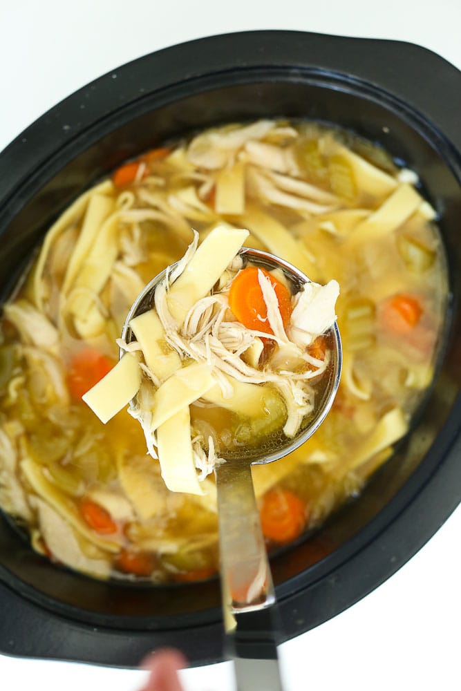 how to make easy crockpot chicken noodle soup recipe ladle with chicken breast, noodles, and carrots coming out of pot
