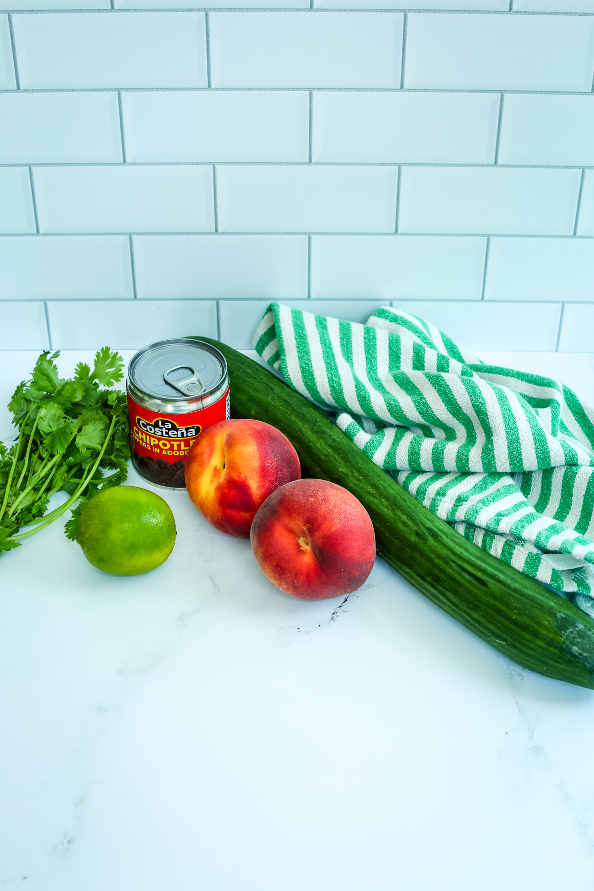 ingredients for peach cucumber salsa, whole cucumber, 2 whole peaches, a can of chipotle peppers, cilantro, and a whole lime with a green and white striped napkin next to the ingredients 
