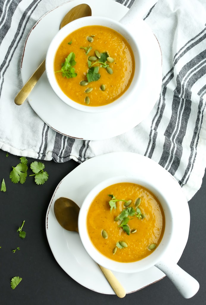 overhead shot of two bowls of butternut squash soup with gold spoons, leafy green garnish, and a white napkin with black stripes underneath