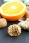 an anti-inflammatory ginger energy ball with a bite taken out of it and a piece of fresh ginger and an orange in the background