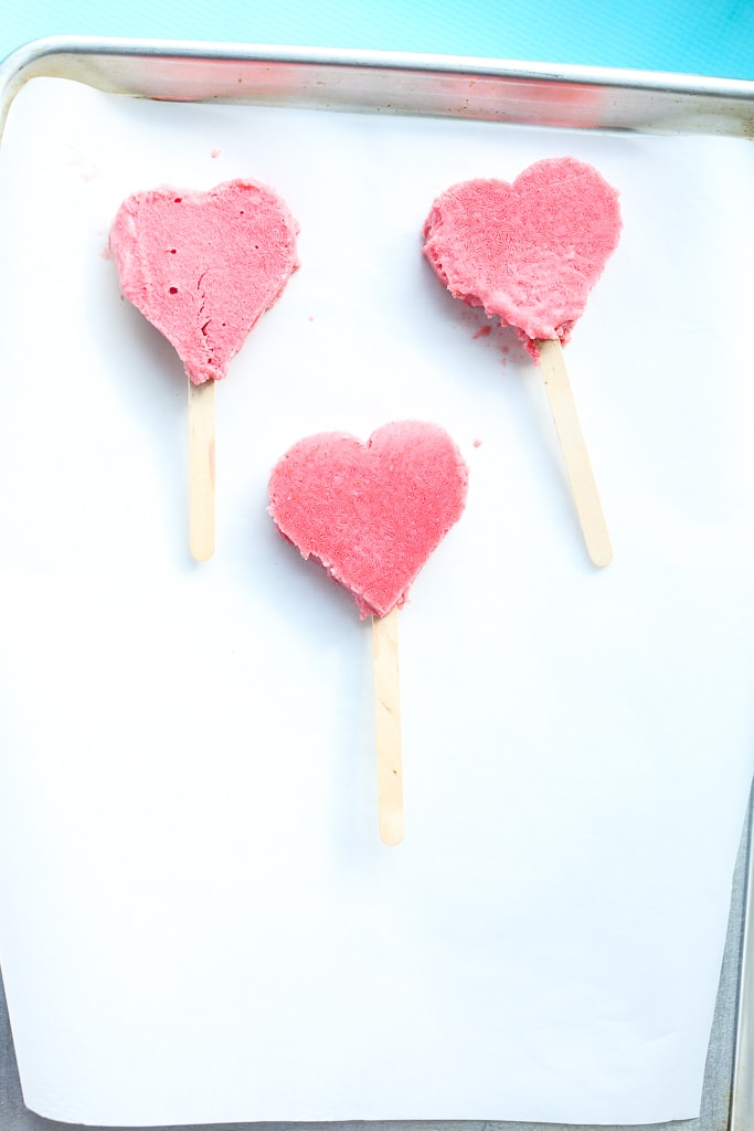 three heart-shaped strawberry bars with wooden sticks on a tray lined with white parchment paper