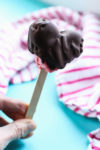 a hand holding a heart-shaped chocolate covered ice cream bar with strawberry ice cream peeking out the bottom. the background is turquoise with a white and pink striped towel.
