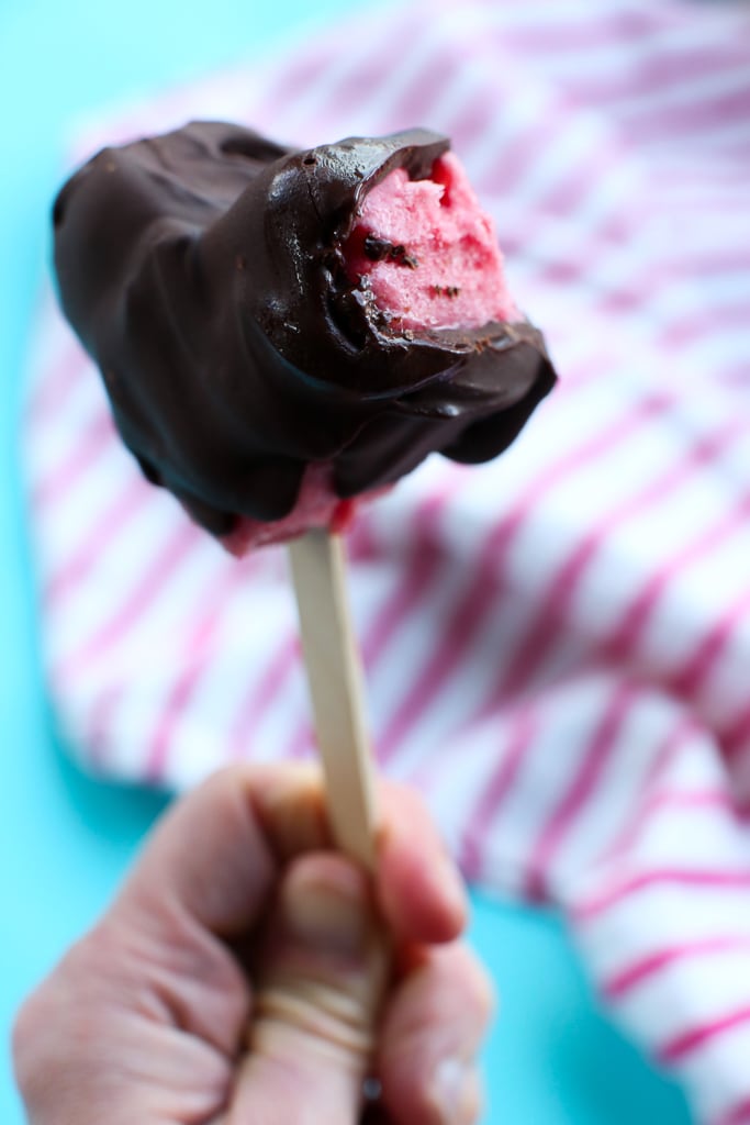 a hand holding a heart-shaped chocolate covered ice cream bar with strawberry ice cream peeking out the bottom. the background is turquoise with a white and pink striped towel.