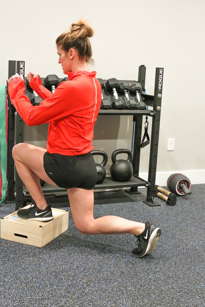 Maryea Flaherty in a red jacket and black shorts completing a reverse lunge exercise with one foot on a brown box