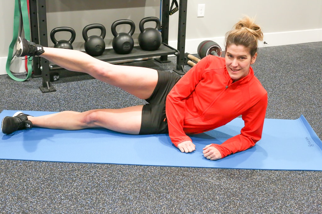 Maryea Flaherty in a red jacket and black shorts laying on her side on a blue mat with one leg extended