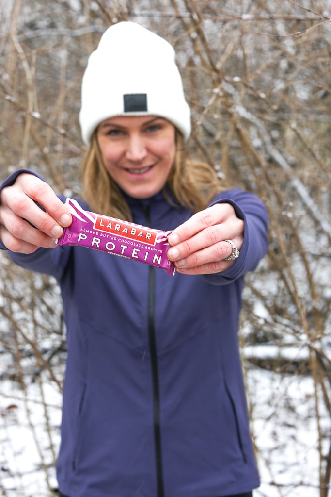 A woman in a purple jacket and white hat holding a Larabar in front of her snowy tree branches in the background