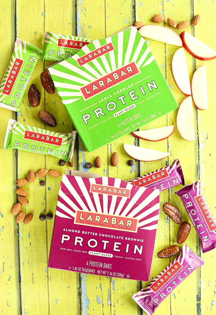 Two boxes of Larabar protein bars with the bars and ingredients coming out of the box. Yellow wood plant background