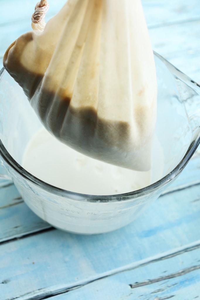 a nut milk bag filled with oats straining into a large glass measuring cup 