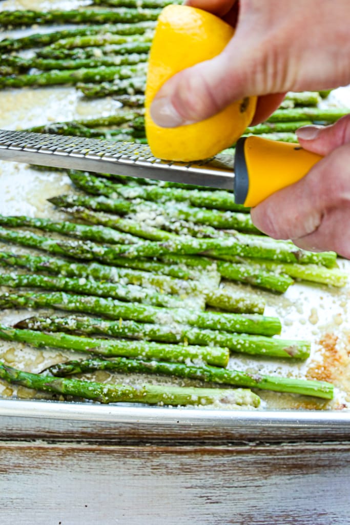 using a zester to add lemon zest to baked asparagus on a baking sheet