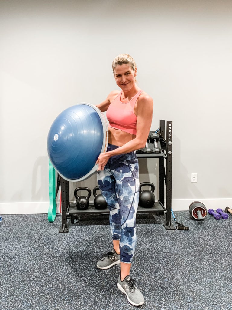 A woman in floral leggings and a sports bra holding a bosu ball in a home gym