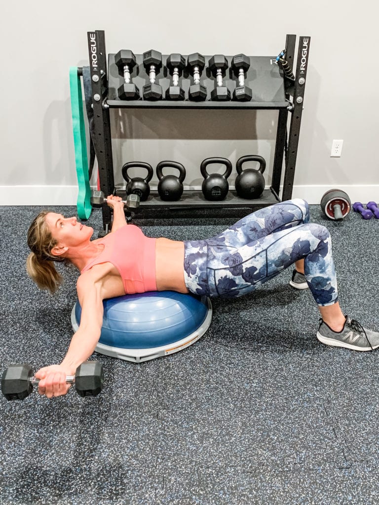 a woman in floral leggings and a sports bra laying on a bosu ball and completing a chest fly exercise with dumbbells
