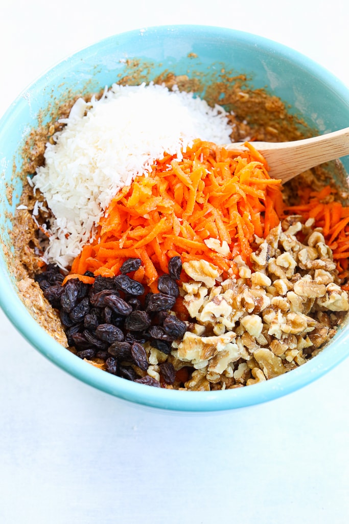 Shredded Carrots, coconut, raisins, and walnuts being folded into batter with a wooden spoon in a turquoise bowl