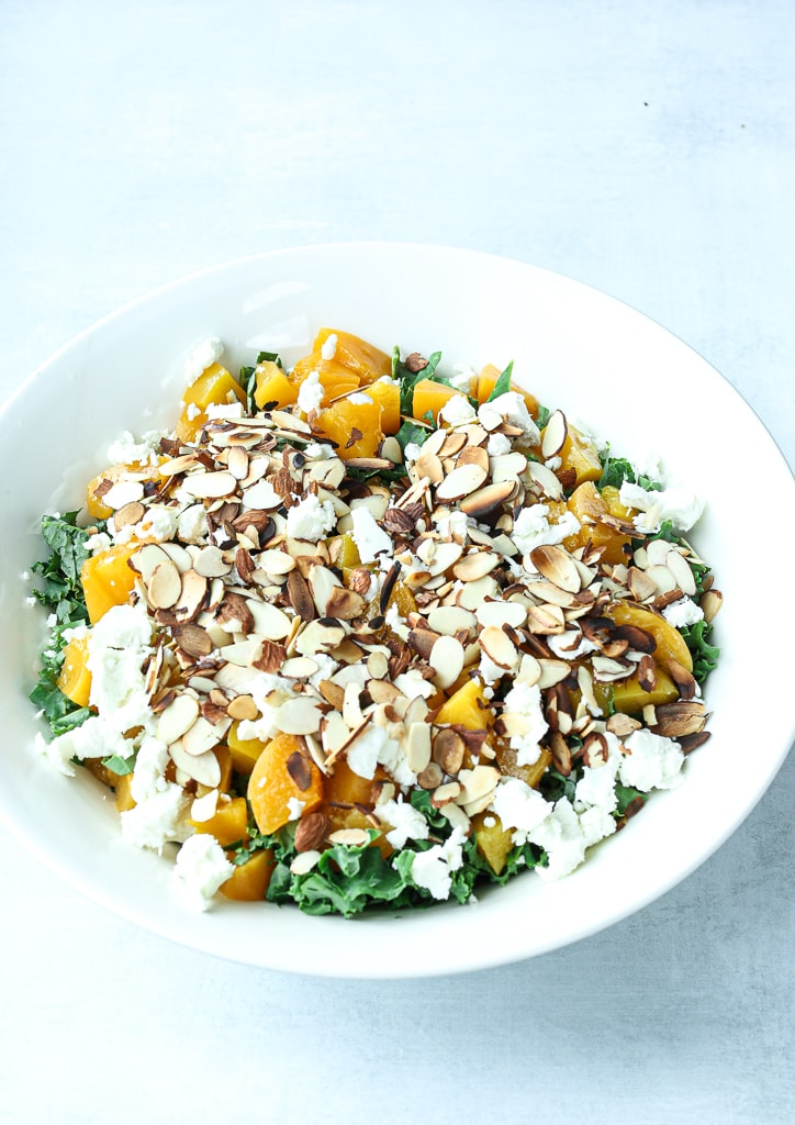 a white bowl with kale, peaches, goat cheese, and sliced almonds