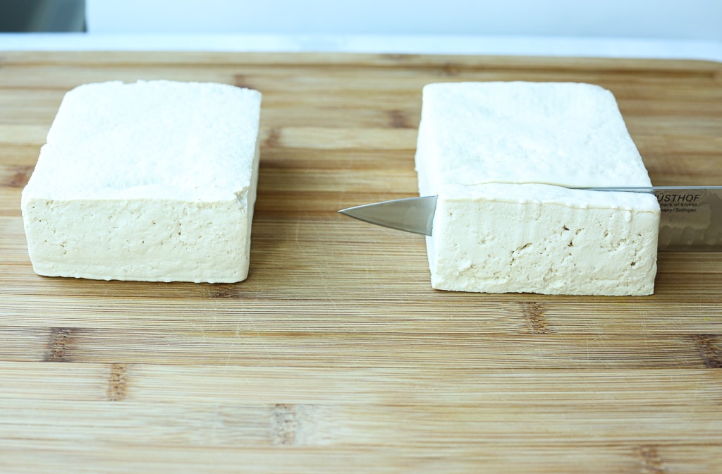two blocks of tofu on a cutting board with a knife slicing one of them