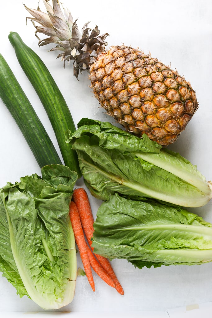 overhead shot of green juice ingredients--pineapple, 2 cucumbers, 3 hearts of romaine, 3 carrots, and a pineapple