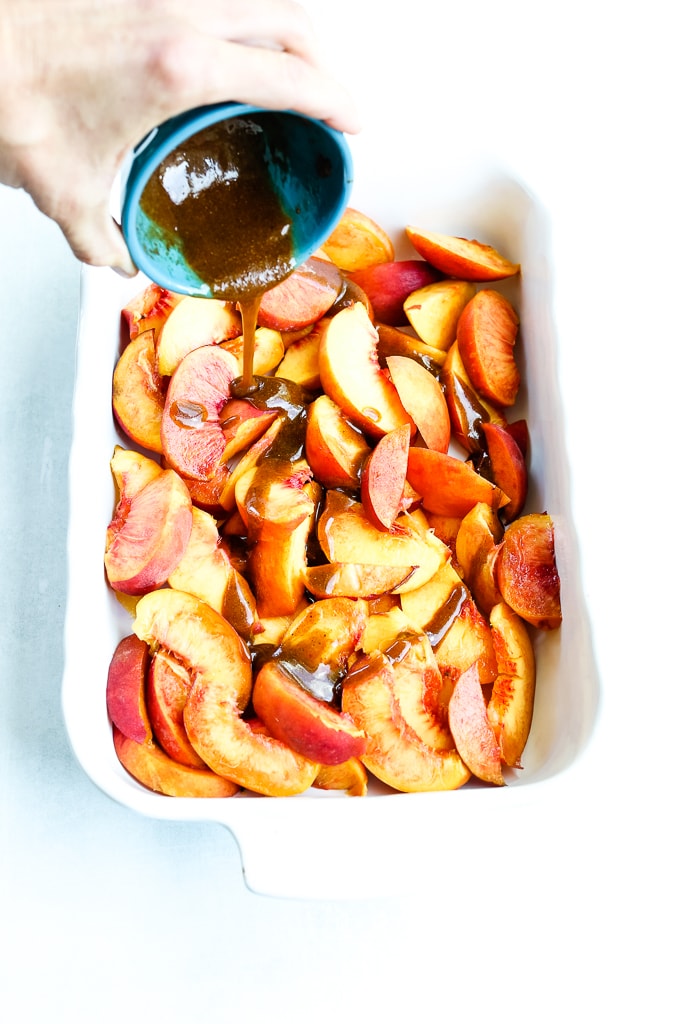 pouring a brown liquid over top of sliced peaches in a white baking dish