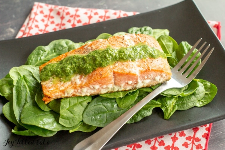 a black plate with salmon and a bed of greens and topped with green pesto sauce