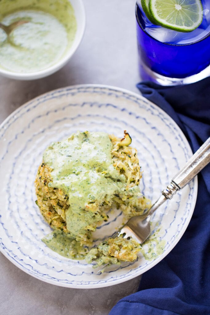 an overhead shot of a zucchini fritter on a white plate with creamy pesto sauce on top