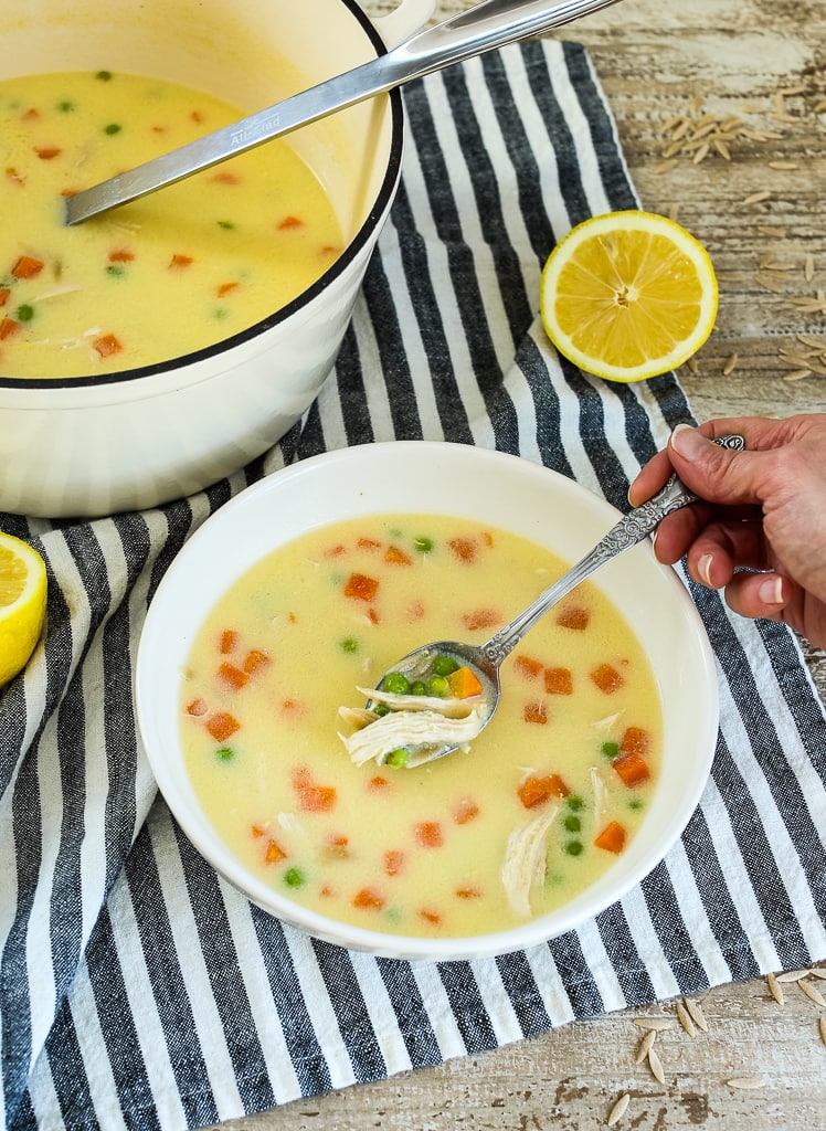 a bowl full of Greek Lemon Chicken Soup with Orzo with a hand and a striped napkin and lemon halves