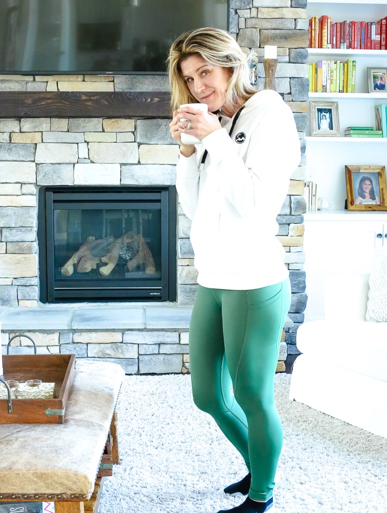 self-care ideas for women: a woman standing with green leggings and a white hooding sipping from a cup of coffee