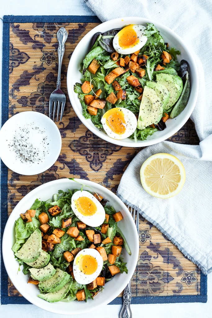 overhead shot of two breakfast salad bowls with a fresh lemon on the table and a small bowl of salt and pepper