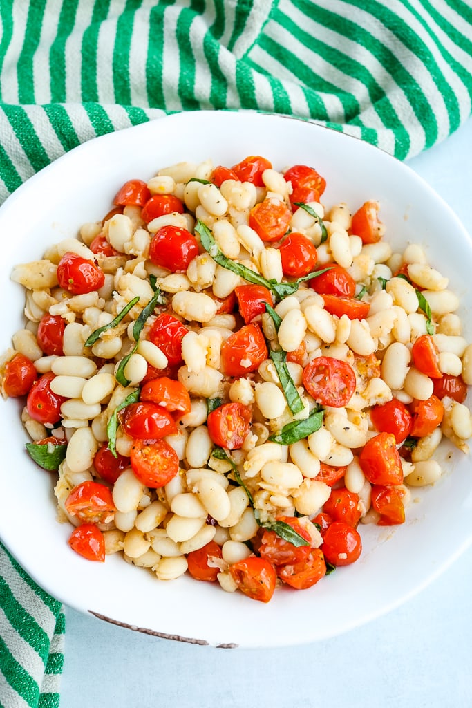 an overhead shot of White Beans with Tomatoes and Fresh Basil in a white bowl with a green and white striped napkin