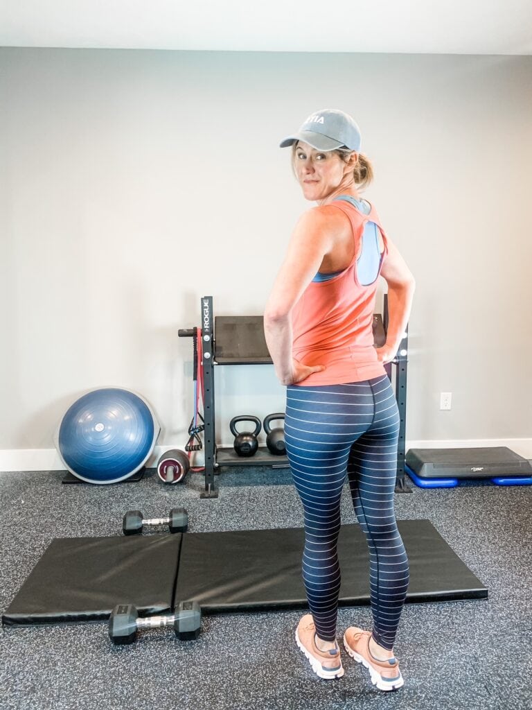 woman standing in a weight room with leggings, tank top, and a hat on