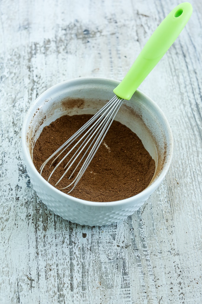 a white bowl with a small whisk with a green handle and brown spices