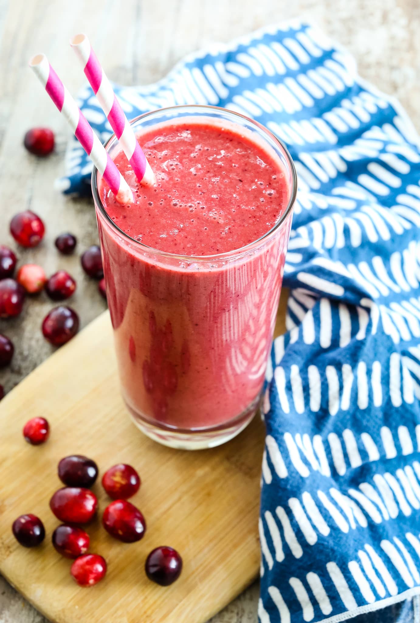 Cranberry Smoothie recipe in a tall glass with two straws and fresh cranberries on the table and a blue and white napkin