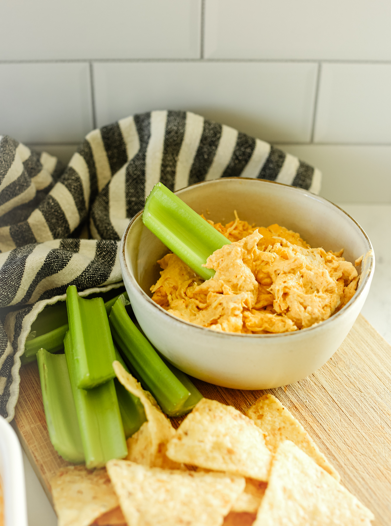 a bowl of orange dip on a wooden tray with celery and chips and a striped napkin