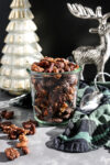 a jar of nuts with a green and black napkin next to them and a silver reindeer and white glass Christmas tree behind the jar