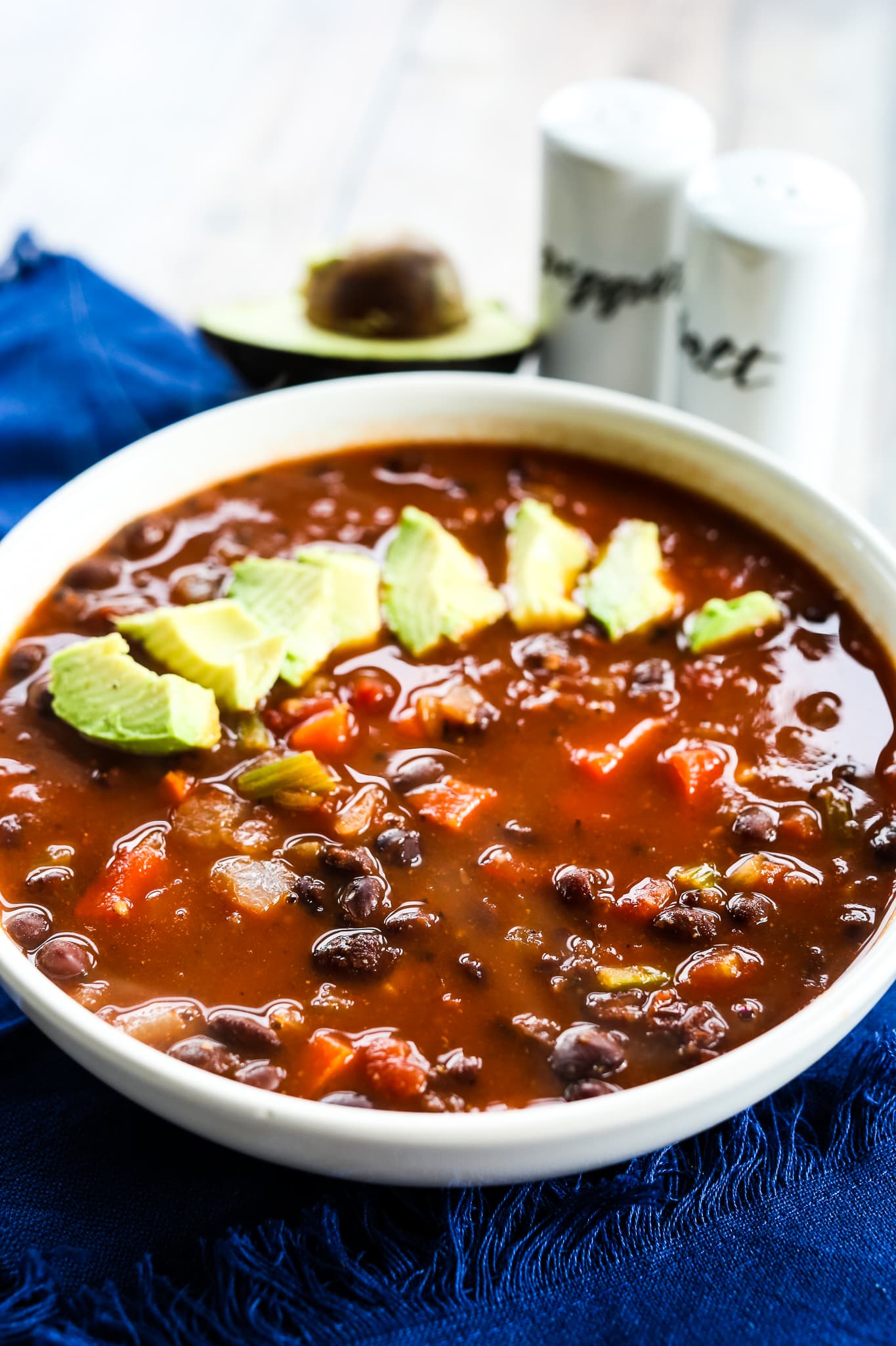 spicy black bean soup with avocado slices on top and salt and pepper in the background and a blue napkin