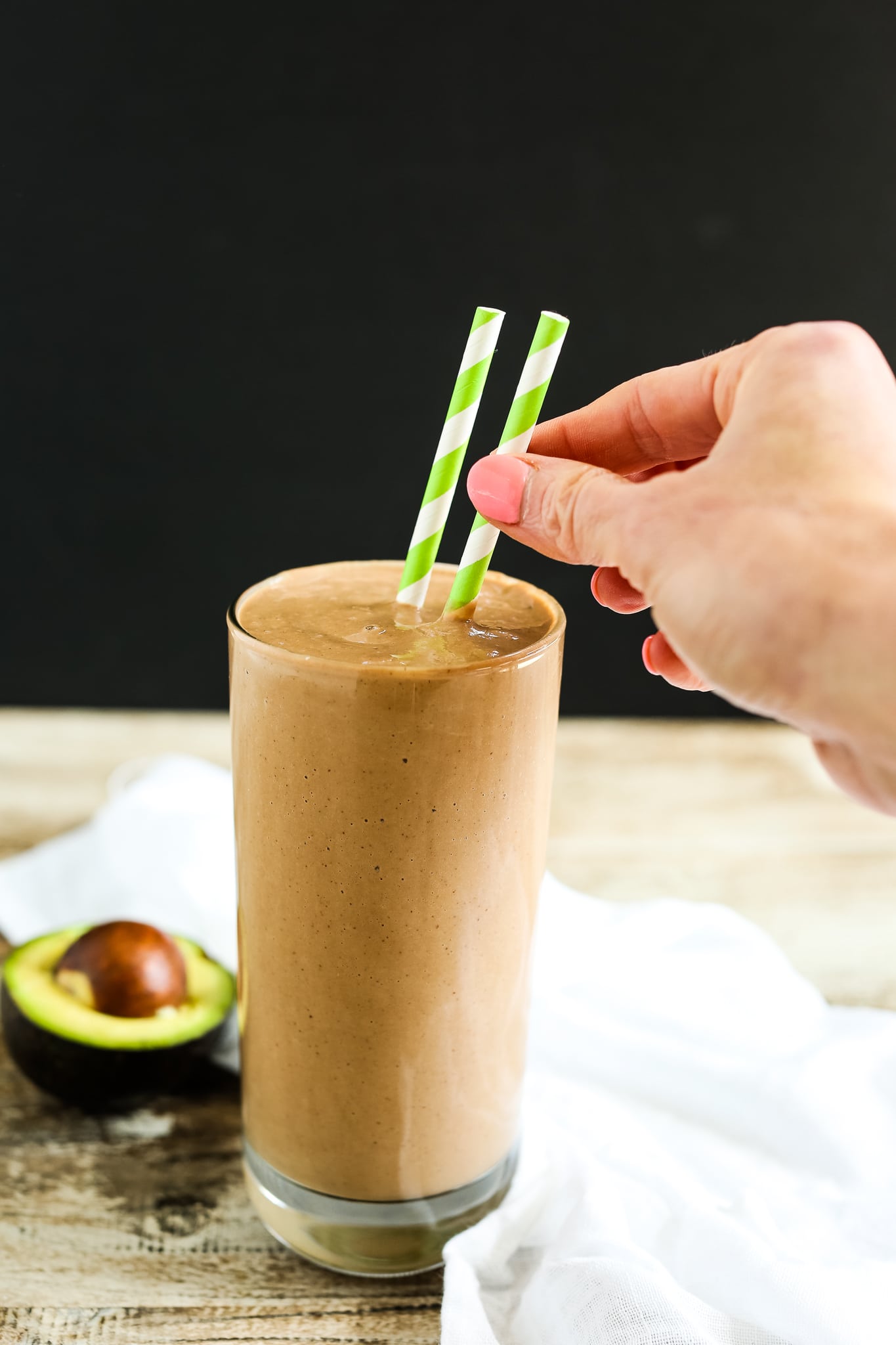 A hand grabbing a green straw that is in a tall glass with a chocolate colored smoothie