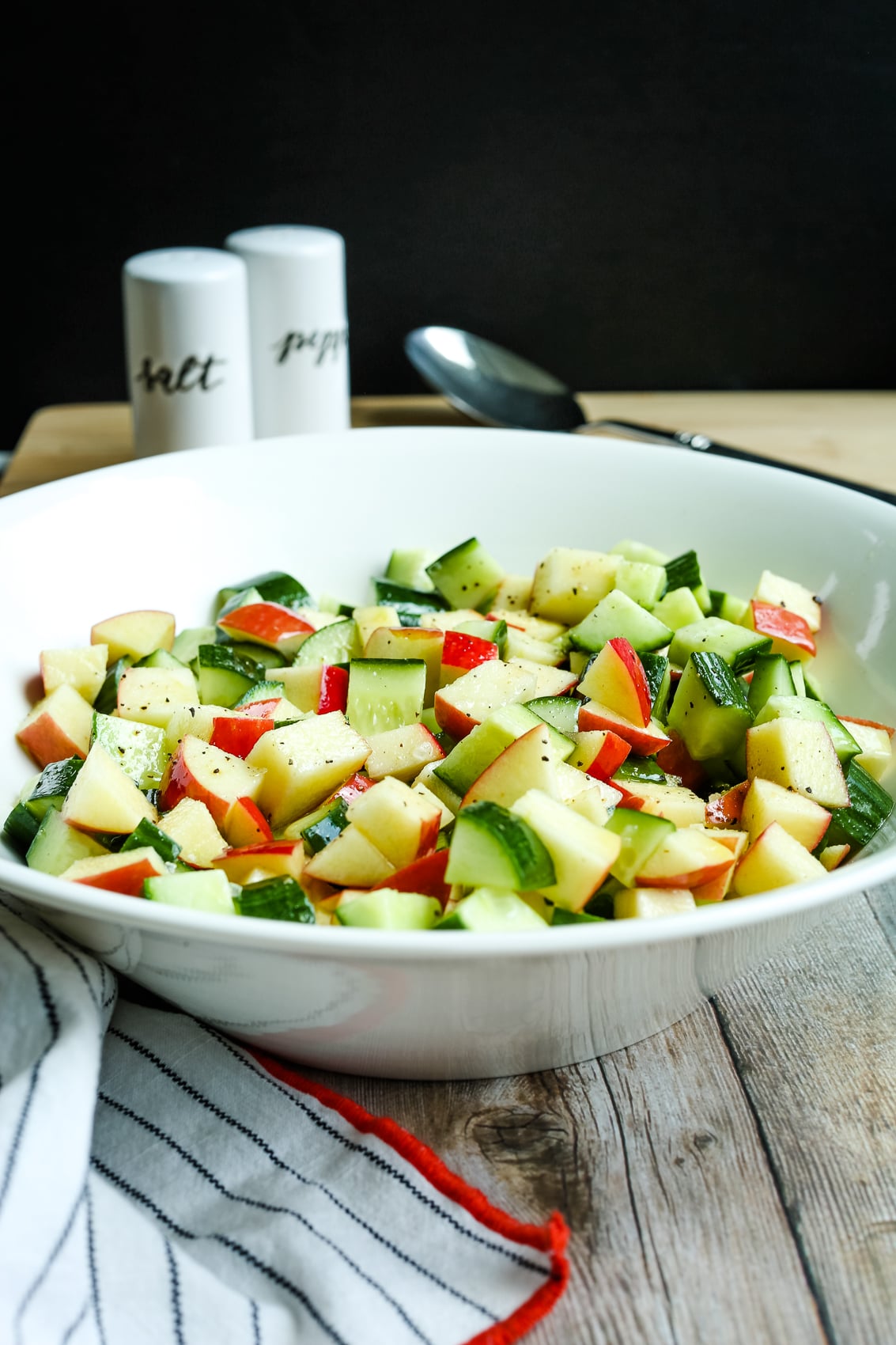 apple cucumber salad in a white bowl with a napkin next to the bowl and salt and pepper shakers in the background.