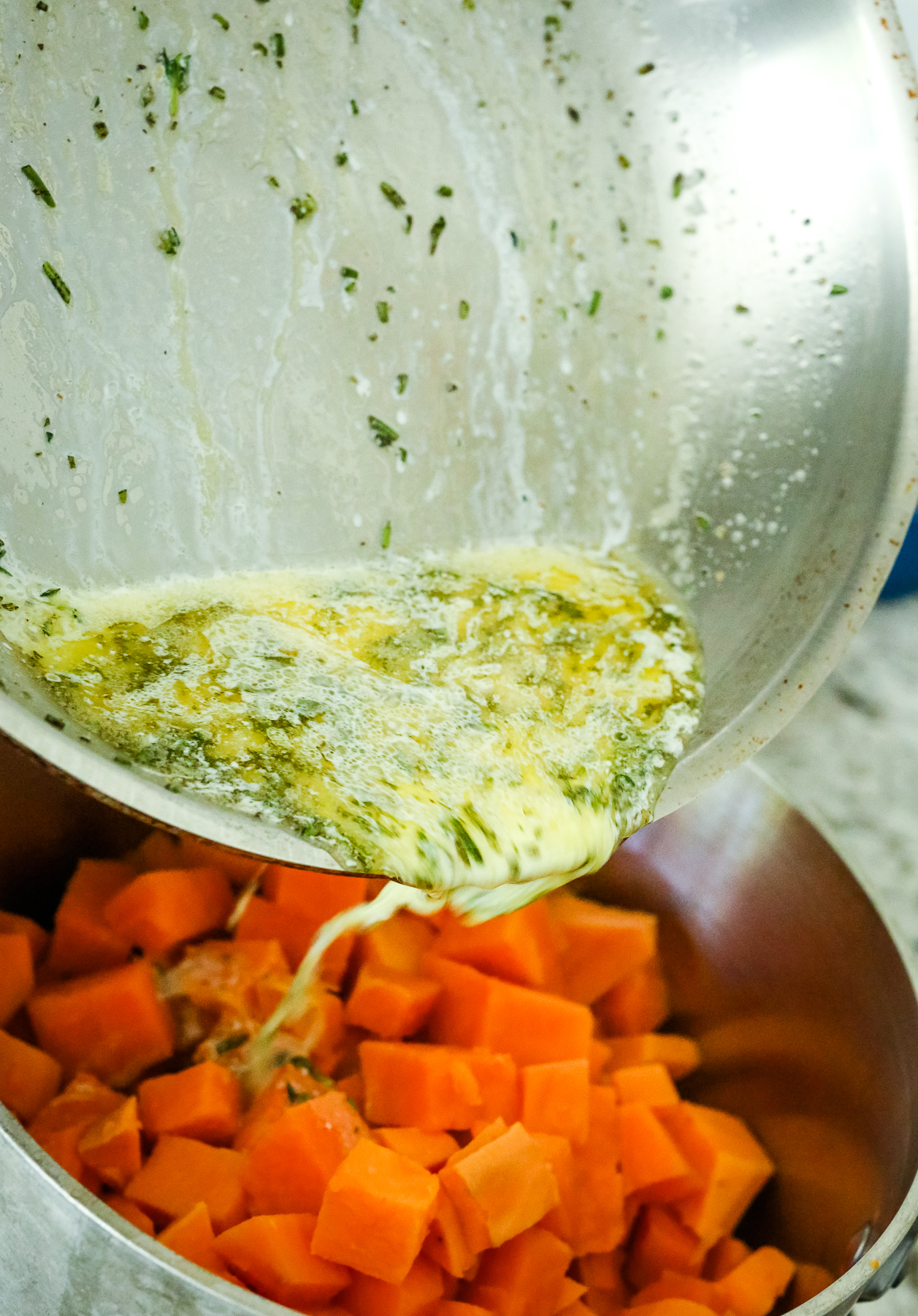 pouring herbed butter from a pan into the pot of steamed sweet potatoes