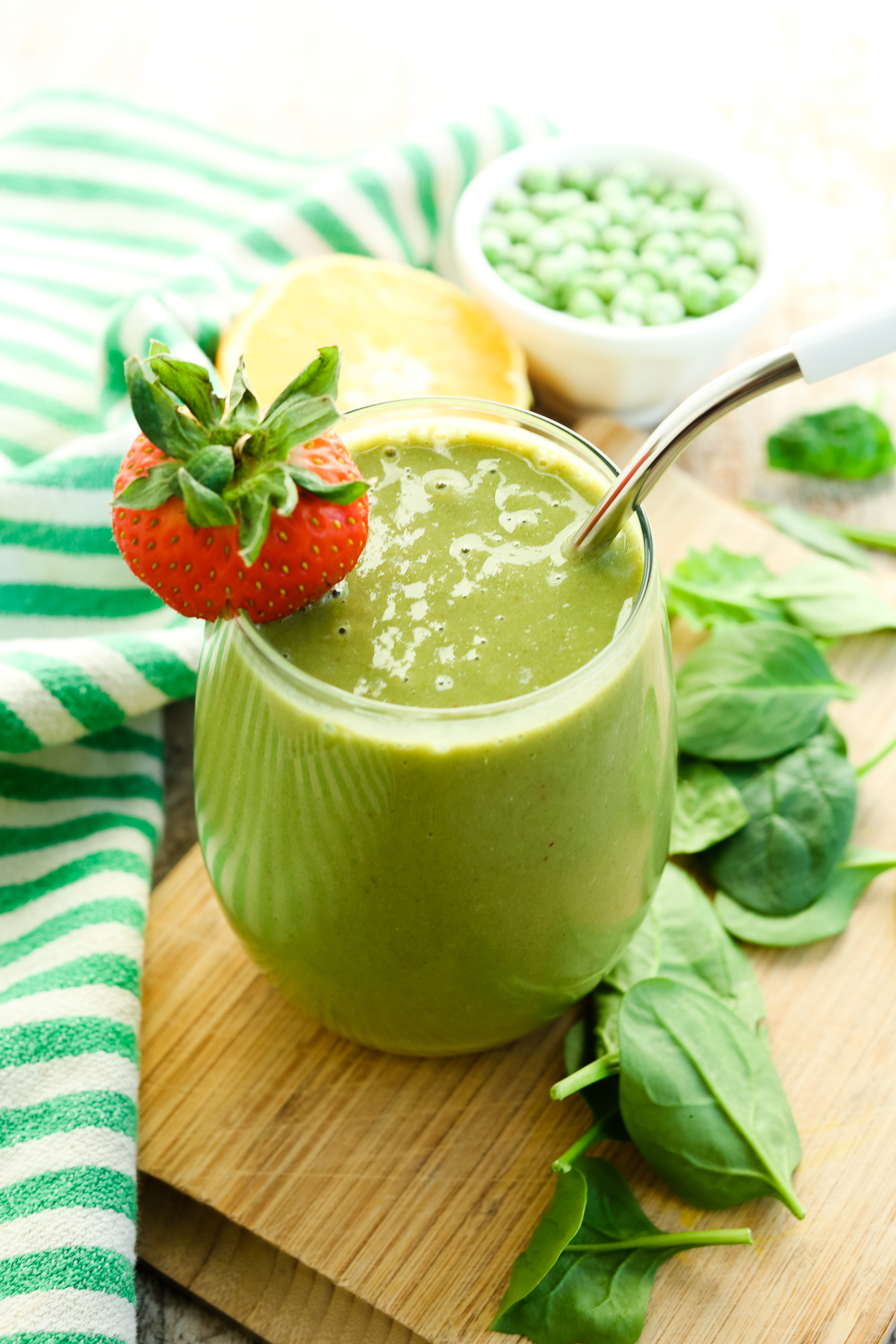 Iron Rich Smoothie in a glass with a stainless steel straw and a strawberry on the rim spinach and orange are in the background along with a green and white striped napkin and a bowl of frozen peas