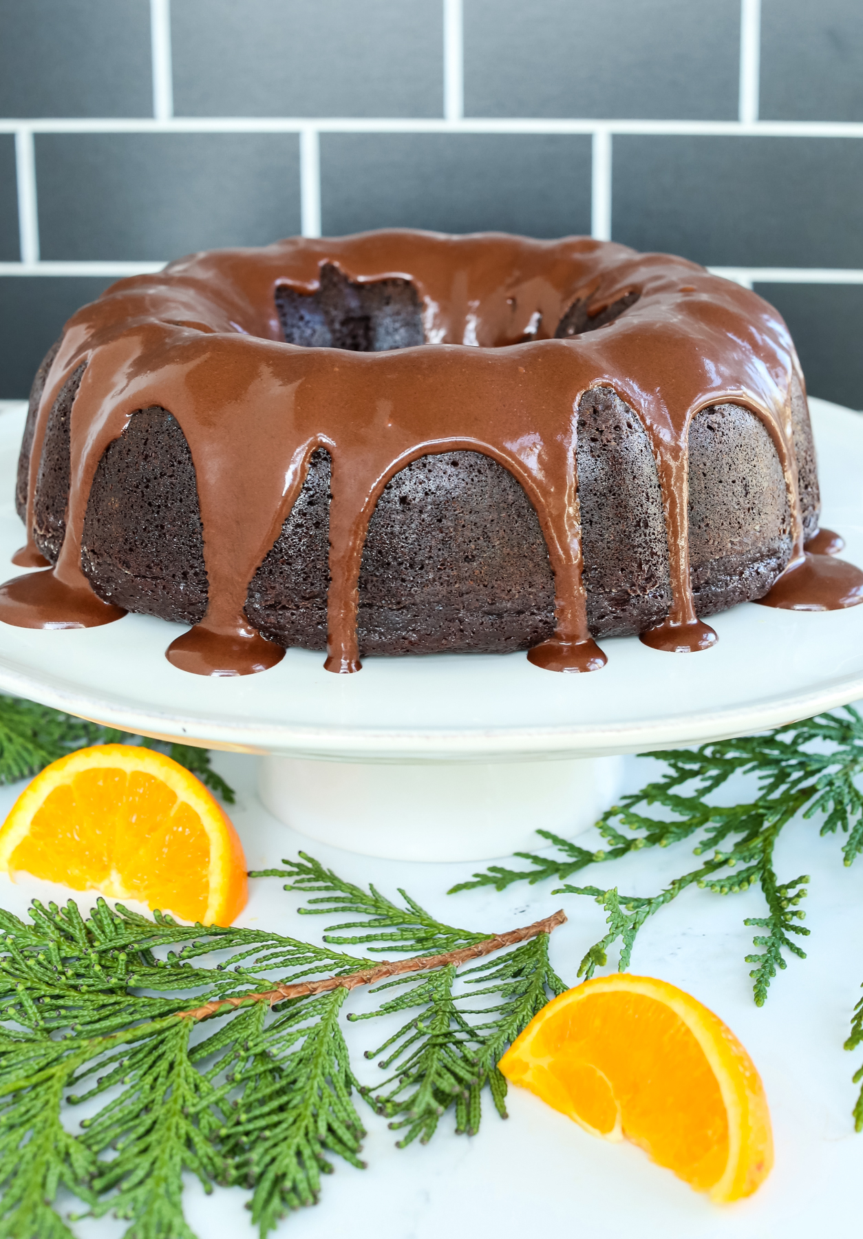 Orange Chocolate Cake on a white stand with chocolate glaze drizzled down the edges. On the table are orange wedges and greenery.