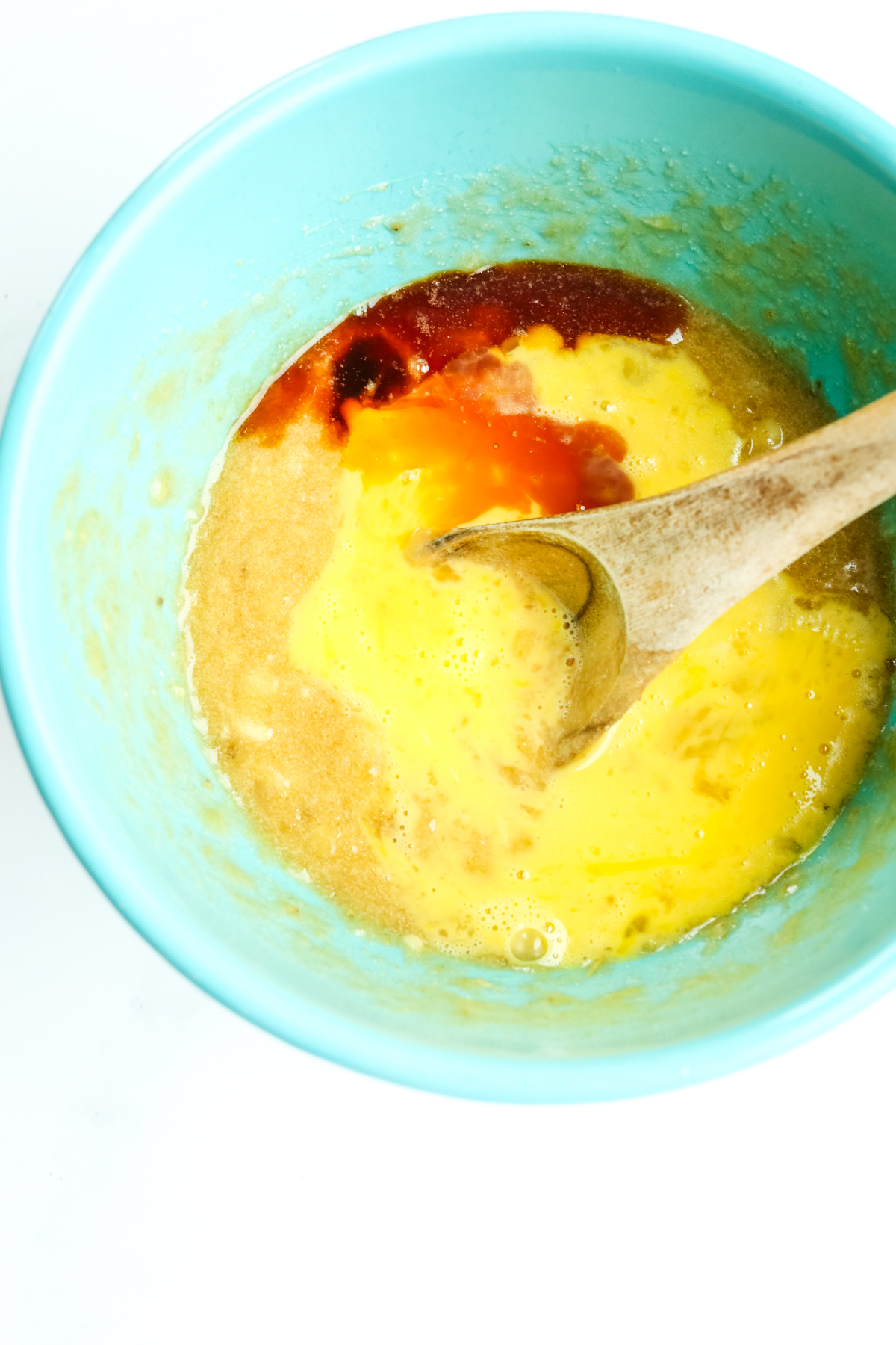 a blue bowl with mashed banana mixture, eggs, and vanilla extract with a wooden spoon preparing to stir them together 