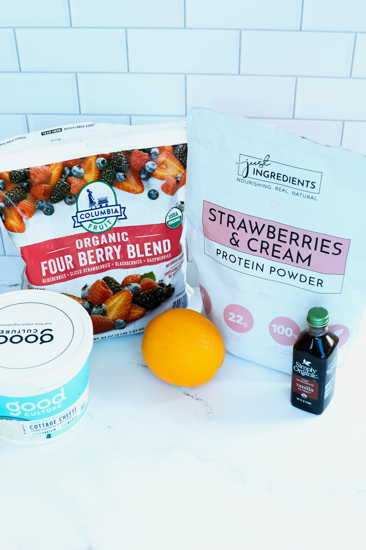 recipe ingredients shown- a bag of protein powder and frozen mixed fruit, an orange, cottage cheese in a plastic tub, and a small bottle of vanilla