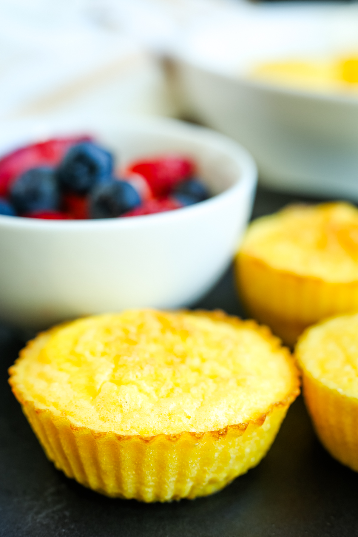 a close up shot of three cottage cheese egg bites on a black plate with a bowl of fruit in the background