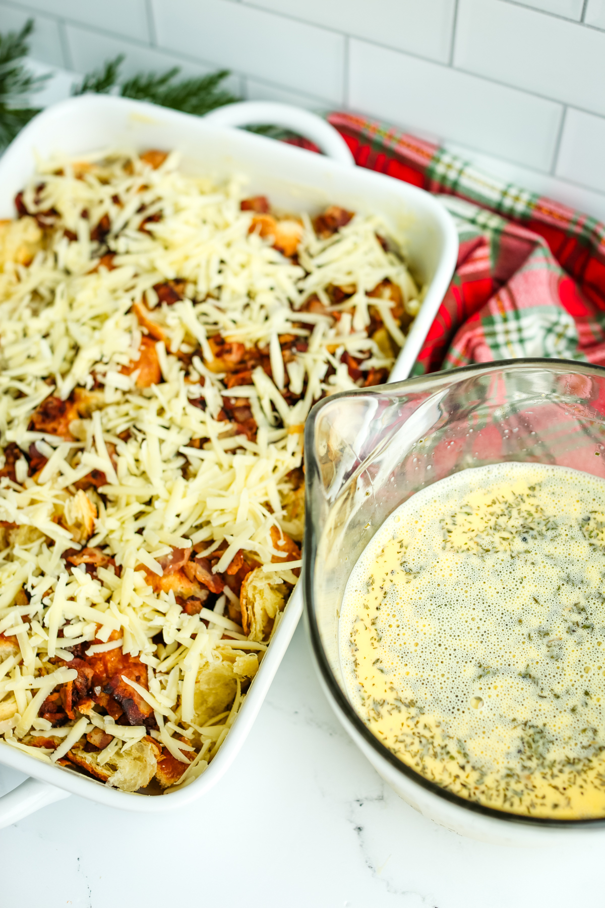 a large liquid measuring cup with eggs, milk, and parsley mixed together next to large baking dish with other ingredients layered in it. 