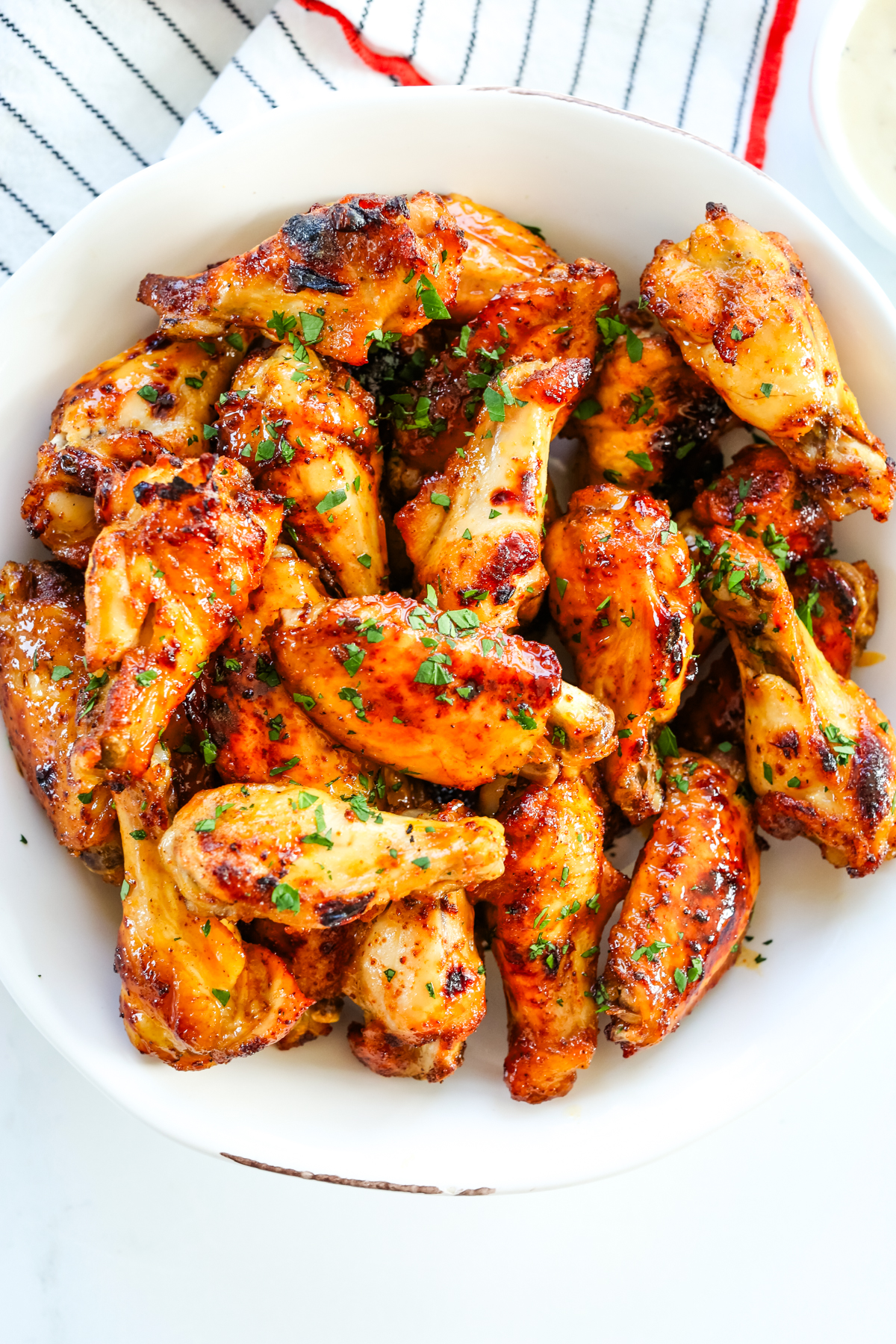 a white bowl on top of a white napkin with black stripes and red stitching, with hot honey chicken wings garnished with fresh parsley 